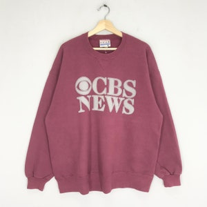 Könnte beinhalten: Ein maroonfarbener Pullover mit einem weißen CBS News-Logo auf der Vorderseite. Das Logo zeigt das CBS-Auge und die Worte "CBS NEWS" in einer fetten, klobigen Schrift.