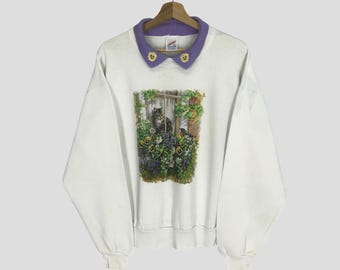 Może przedstawiać: Biała bluza z fioletowym kołnierzem i złotymi guzikami. Bluza ma nadrukowany obraz kota siedzącego w ogrodzie z kwiatami.