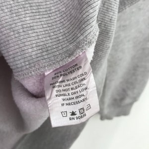 May include: A white tag with black text detailing the fabric content and care instructions for a gray sweatshirt. The tag reads "80% Cotton 20% Polyester", "Machine wash cold with like colors", "Do not bleach", "Tumble dry low", "Warm iron", "If necessary", and "RN 90836".