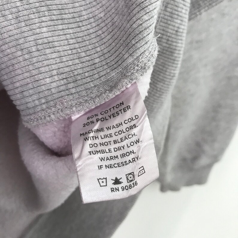 May include: A white tag with black text detailing the fabric content and care instructions for a gray sweatshirt. The tag reads "80% Cotton 20% Polyester", "Machine wash cold with like colors", "Do not bleach", "Tumble dry low", "Warm iron", "If necessary", and "RN 90836".