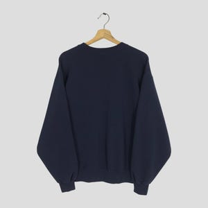May include: A navy blue sweatshirt hanging on a wooden hanger. The sweatshirt has long sleeves and a crew neck. The cuffs and hem are ribbed. The background is a solid light gray.