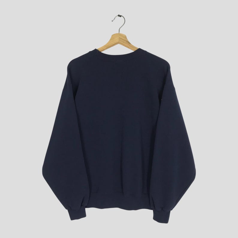 May include: A navy blue sweatshirt hanging on a wooden hanger. The sweatshirt has long sleeves and a crew neck. The cuffs and hem are ribbed. The background is a solid light gray.