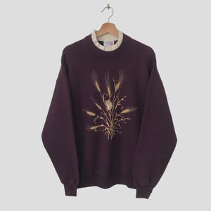Puede incluir: Una sudadera de color morado oscuro con un diseño bordado en dorado de tallos de trigo y una flor en la parte delantera. La sudadera tiene cuello redondo y mangas largas.
