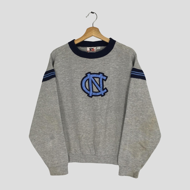 K&ouml;nnte beinhalten: Ein graues Sweatshirt mit einem blau-hellblauen Logo auf der Vorderseite. Das Sweatshirt hat einen marineblauen Kragen und blaue Streifen auf den &Auml;rmeln. Es h&auml;ngt an einem Holzb&uuml;gel.