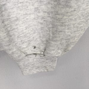 May include: Close-up of a light gray sweatshirt sleeve. The fabric has a textured appearance with a ribbed cuff. There are several small dark spots on the fabric. The background is a plain, light gray.