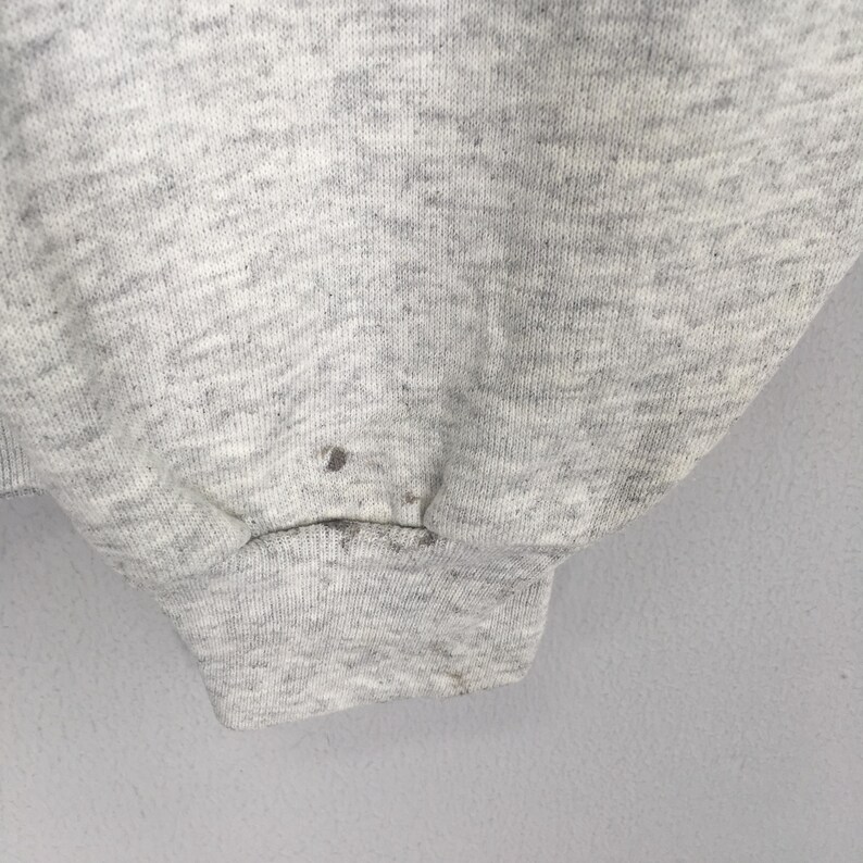 May include: Close-up of a light gray sweatshirt sleeve. The fabric has a textured appearance with a ribbed cuff. There are several small dark spots on the fabric. The background is a plain, light gray.