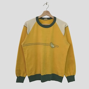 Könnte beinhalten: Ein gelbes Langarm-Sweatshirt mit cremefarbenen Schulterpartien und dunkelgrünen Zierleisten. Das Sweatshirt hat ein horizontales Streifendesign mit einem Logo auf der Vorderseite. Das Kleidungsstück hängt an einem Holzbügel.