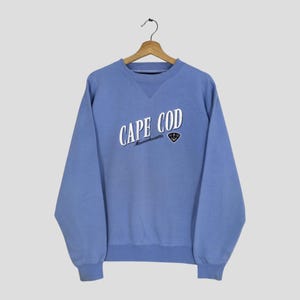 Op de afbeelding: Een lichtblauw sweatshirt met ronde hals en de witte tekst CAPE COD Massachusetts op de borst gedrukt. Onder de tekst staat een klein zwart driehoekig embleem met een witte ankergrafiek. Het kledingstuk heeft lange mouwen en boorden.