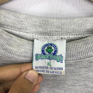 May include: Gray sweatshirt with a tag that says "Official Wear Se&ntilde;or Frog's" in a circle with a green frog inside. The tag also says "XL" and "50% Polyester / 50% Algodon Hecho en Mexico".