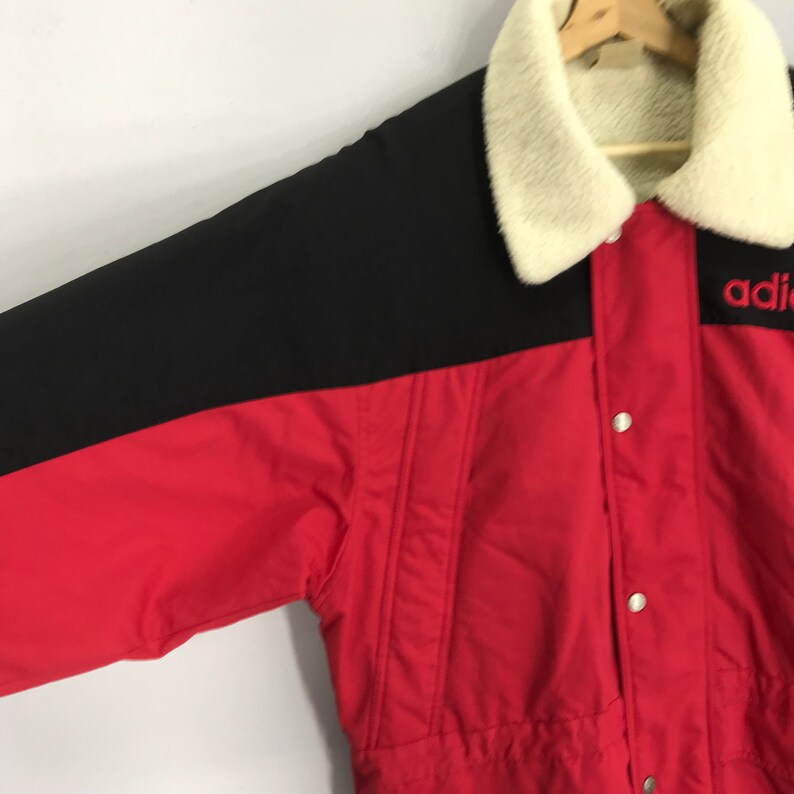 May include: A red and black jacket with a white faux fur collar. The jacket has a button closure and the word "adidas" is embroidered on the right side of the collar in red.