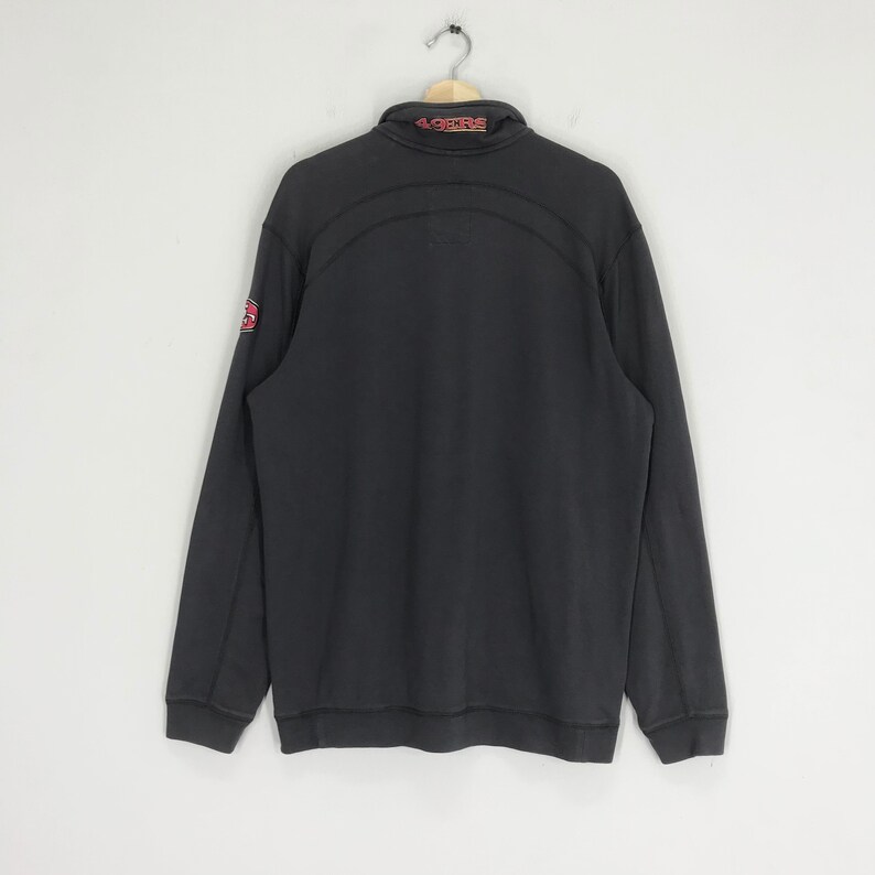 Peut inclure: Sweat-shirt &agrave; manches longues noir avec un logo rouge et blanc sur la manche gauche et "49ERS" en rouge &agrave; l'arri&egrave;re du col.