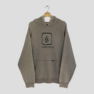 May include: A tan hoodie with a front pocket and a hood. The hoodie features a black square logo with the word "volcom" printed below it. The hoodie is hanging on a wooden hanger.