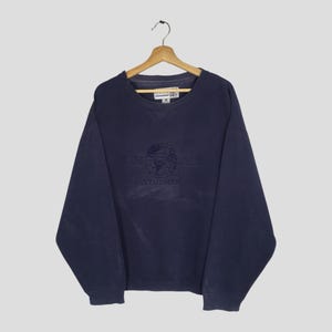 May include: Navy blue Universal Studios sweatshirt with the logo embroidered on the front. The long-sleeved crew neck sweatshirt is hanging on a wooden hanger. The sweatshirt is a solid color.
