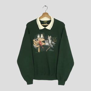 Puede incluir: Sudadera verde oscuro con cuello color crema. La parte delantera presenta un gráfico de pájaros, gatos y una pajarera. El texto "Bird Watchers" está impreso debajo. La sudadera tiene mangas largas y un corte holgado.
