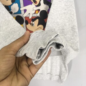 May include: A light gray sweatshirt featuring a colorful graphic of Mickey Mouse in various poses. The graphic is set against a backdrop of red, blue, and purple squares. The sleeve cuff is visible.