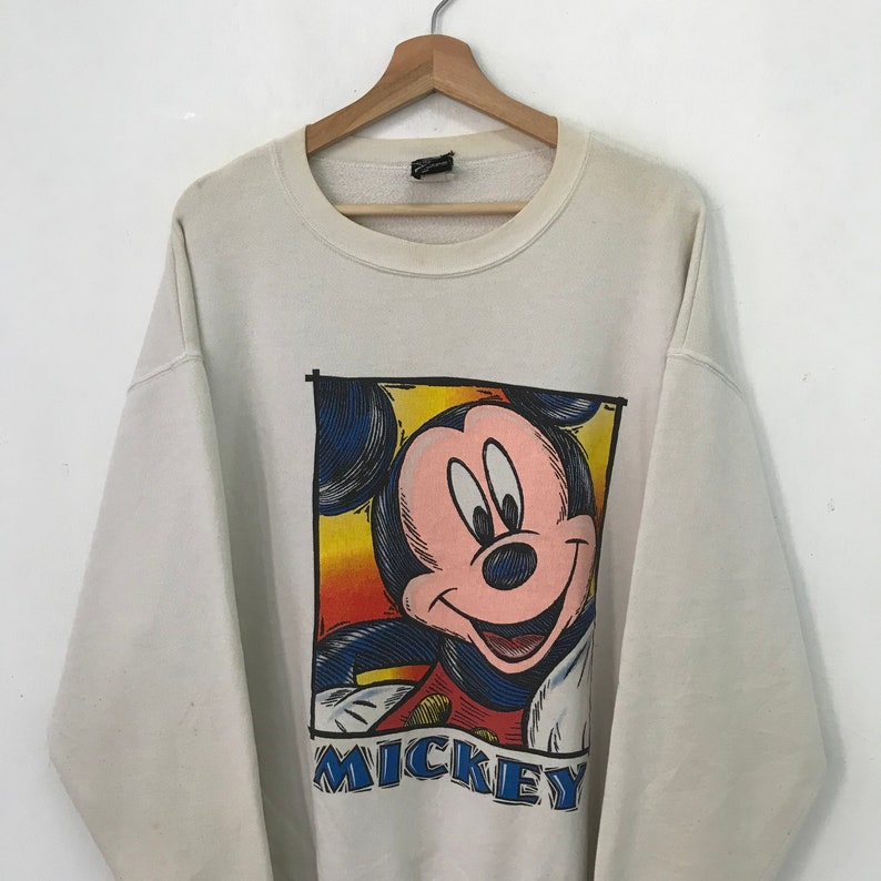 May include: White sweatshirt with a colorful cartoon image of Mickey Mouse. The image is framed in a black outline and the word "MICKEY" is printed below the image.
