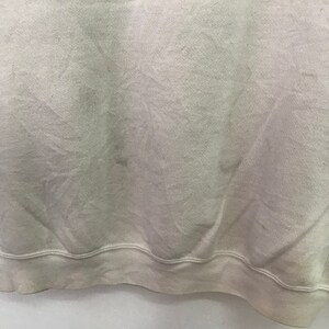 May include: A close-up of the bottom hem of a light beige sweatshirt. The fabric is soft and has a slight texture. The hem is folded over and stitched.
