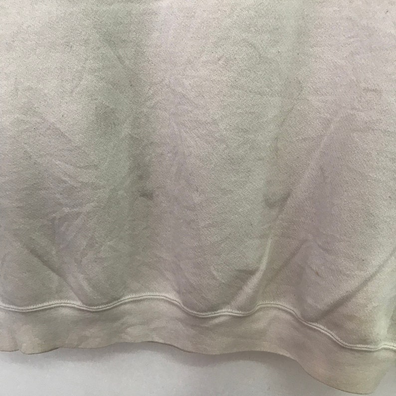 May include: A close-up of the bottom hem of a light beige sweatshirt. The fabric is soft and has a slight texture. The hem is folded over and stitched.