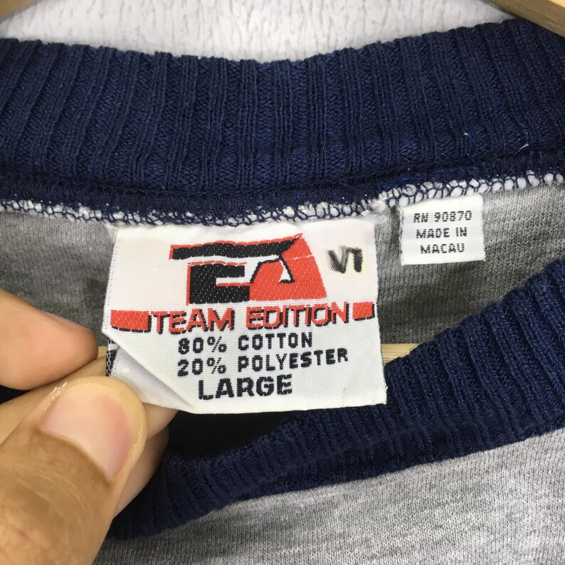 K&ouml;nnte beinhalten: Nahaufnahme eines grau-blauen Ringer-T-Shirts mit Etikett. Das Etikett lautet "TEAM EDITION" und gibt die Stoffzusammensetzung mit 80 % Baumwolle und 20 % Polyester an. Die Gr&ouml;&szlig;e ist "LARGE". Das Shirt wurde in Macau hergestellt.