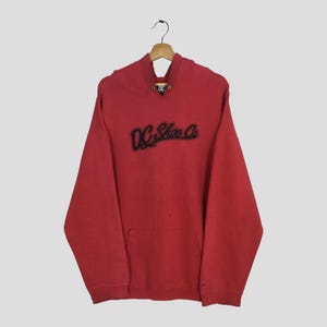 May include: Red hoodie with the text "DC Shoe Co" in black script across the chest. The hoodie has a front pocket and a hood with a drawstring. The garment is hanging on a wooden hanger.