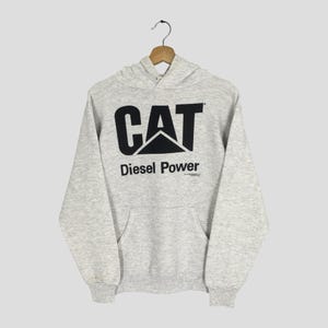 Puede incluir: Una sudadera con capucha de manga larga en color gris jaspeado claro que presenta un gran logotipo negro de CAT y el texto Diesel Power impreso en el frente. La prenda incluye un bolsillo tipo canguro, pu&ntilde;os acanalados y un dobladillo acanalado.