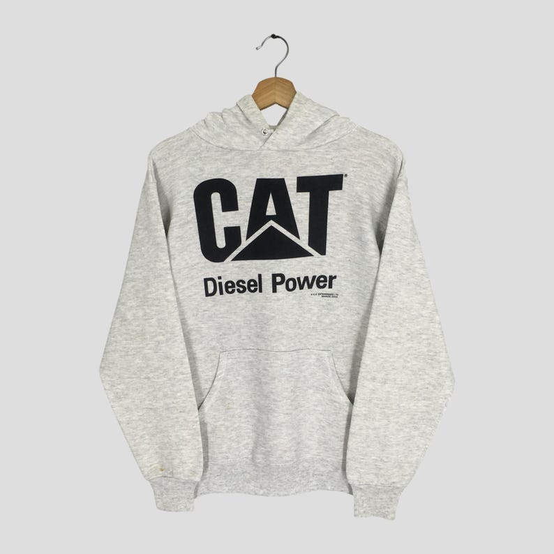 Puede incluir: Una sudadera con capucha de manga larga en color gris jaspeado claro que presenta un gran logotipo negro de CAT y el texto Diesel Power impreso en el frente. La prenda incluye un bolsillo tipo canguro, pu&ntilde;os acanalados y un dobladillo acanalado.