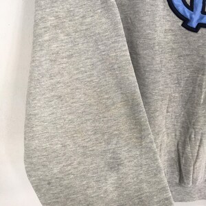 K&ouml;nnte beinhalten: Ein graues Sweatshirt mit einem hellblauen und marineblauen Emblem auf der oberen rechten Seite. Das Sweatshirt besteht aus einem weichen Material und hat einen Rundhalsausschnitt. Der &Auml;rmel ist im Bild sichtbar.