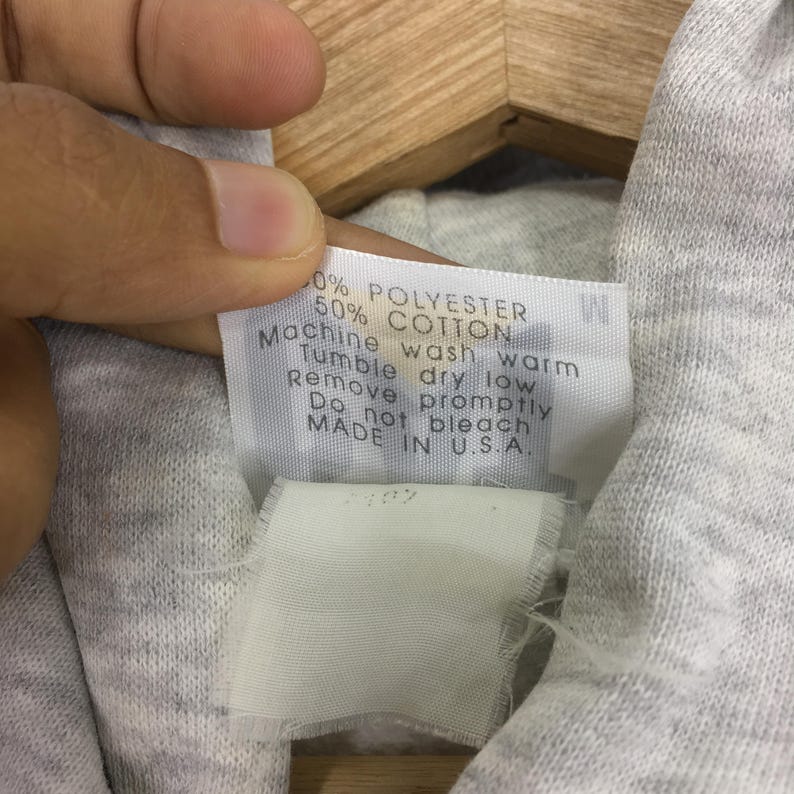 Puede incluir: Una etiqueta de cuidado de tela blanca unida a un textil gris jaspeado claro. La etiqueta presenta texto negro que dice 50% POLYESTER 50% COTTON, Machine wash warm, Tumble dry low, Remove promptly, Do not bleach, MADE IN U.S.A. y la talla M.