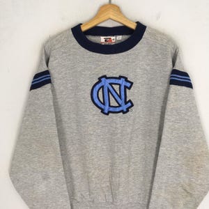 K&ouml;nnte beinhalten: Ein graues Sweatshirt mit einem blau-hellblauen Logo. Das Sweatshirt hat einen blauen Kragen und blaue Streifen auf den &Auml;rmeln. Das Logo ist ein stilisiertes "NC" in Hellblau.