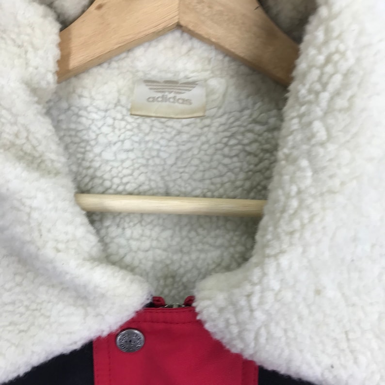 May include: A red and black jacket with a white sherpa lining and collar. The jacket has a zipper closure and a button-up flap. The label on the inside of the jacket reads "adidas".