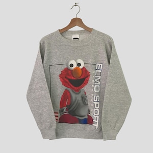 Könnte beinhalten: Ein graues Sweatshirt mit einer Grafik von Elmo aus der Sesamstraße in einem Sport-Design. Elmo trägt eine graue Weste und rote Shorts, mit dem Schriftzug "ELMO SPORT" vertikal auf der rechten Seite.