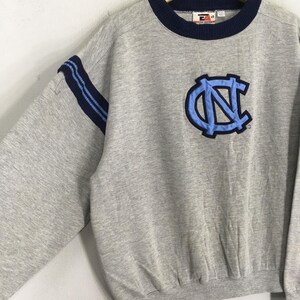 K&ouml;nnte beinhalten: Ein graues Sweatshirt mit marineblauem Kragen und &Auml;rmelstreifen. Auf der Vorderseite befindet sich ein hellblaues und marineblaues Emblem. Das Sweatshirt ist im Rundhals-Stil.