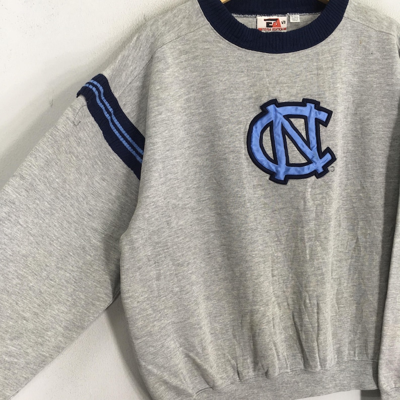 K&ouml;nnte beinhalten: Ein graues Sweatshirt mit marineblauem Kragen und &Auml;rmelstreifen. Auf der Vorderseite befindet sich ein hellblaues und marineblaues Emblem. Das Sweatshirt ist im Rundhals-Stil.