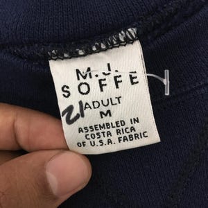 May include: Close-up of a navy blue garment tag. The tag reads "M.J. SOFFE" with "ADULT M" and "ASSEMBLED IN COSTA RICA OF U.S.A. FABRIC" printed on it. A handwritten "2" is also visible.