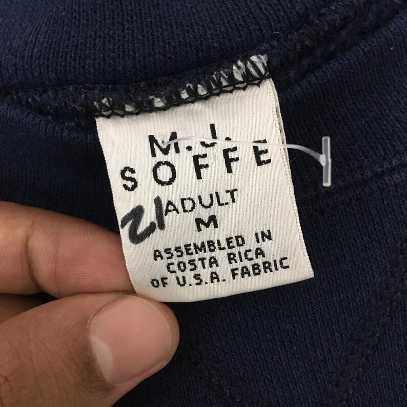 May include: Close-up of a navy blue garment tag. The tag reads "M.J. SOFFE" with "ADULT M" and "ASSEMBLED IN COSTA RICA OF U.S.A. FABRIC" printed on it. A handwritten "2" is also visible.
