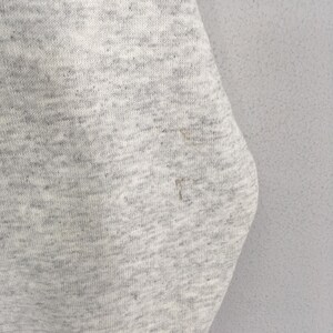May include: Close-up of a light gray fabric with a speckled texture. The fabric has a slightly worn appearance with a few small stains. The material appears to be a knit or woven textile, possibly a sweatshirt or similar garment.