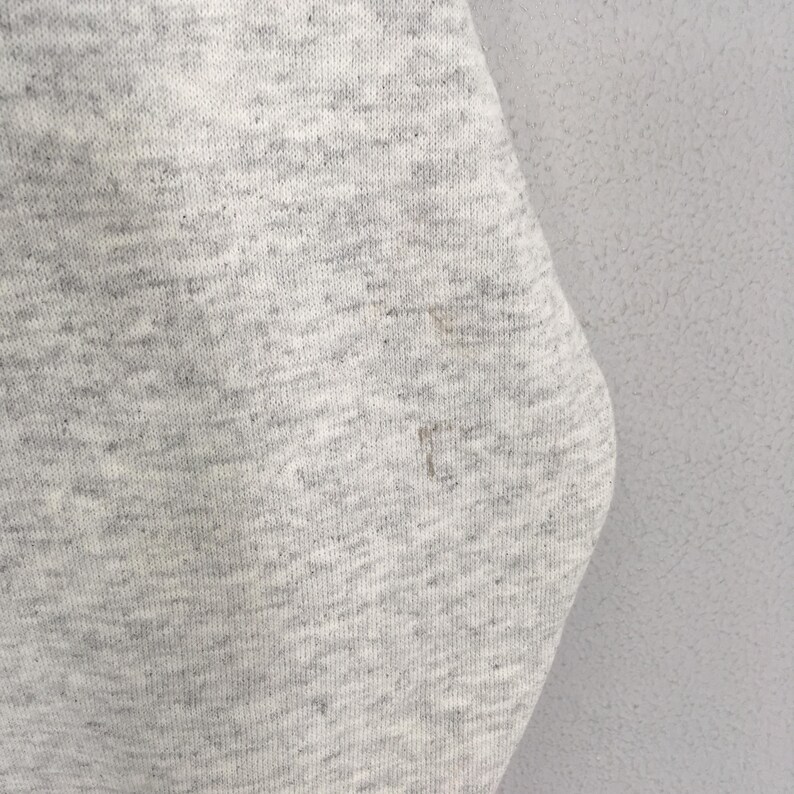 May include: Close-up of a light gray fabric with a speckled texture. The fabric has a slightly worn appearance with a few small stains. The material appears to be a knit or woven textile, possibly a sweatshirt or similar garment.