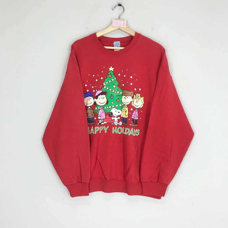 May include: Red sweatshirt with a Christmas tree and the Peanuts characters, including Snoopy, Charlie Brown, Lucy, Linus, and Sally. The text "HAPPY HOLIDAYS" is printed below the characters.
