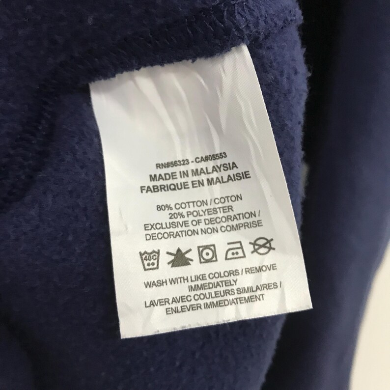 May include: A white tag with black text detailing the care instructions for a navy blue sweatshirt. The tag includes the RN and CA numbers, the country of origin, fabric content, and washing instructions. The text reads: "RN#56323 - CA#05553 MADE IN MALAYSIA FABRIQUE EN MALAISIE 80% COTTON/COTON 20% POLYESTER EXCLUSIVE OF DECORATION/DECORATION NON COMPRISE 40C ☆巴回風 WASH WITH LIKE COLORS/REMOVE IMMEDIATELY LAVER AVEC COULEURS SIMILAIRES/ENLEVER IMMEDIATEMENT"