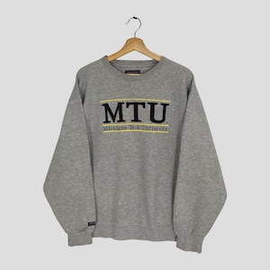 Könnte beinhalten: Ein graues Sweatshirt mit den schwarzen Buchstaben "MTU", einem gelben Streifen darüber und den Worten "Michigan Tech University" darunter. Das Sweatshirt hängt an einem Holzbügel.