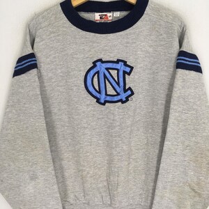 K&ouml;nnte beinhalten: Ein graues Sweatshirt mit marineblauem Kragen und &Auml;rmelstreifen. Auf der Vorderseite befindet sich ein hellblaues und marineblaues Logo. Das Sweatshirt hat lange &Auml;rmel und einen gerippten Saum. Das Logo ist ein stilisiertes "NC"-Design.
