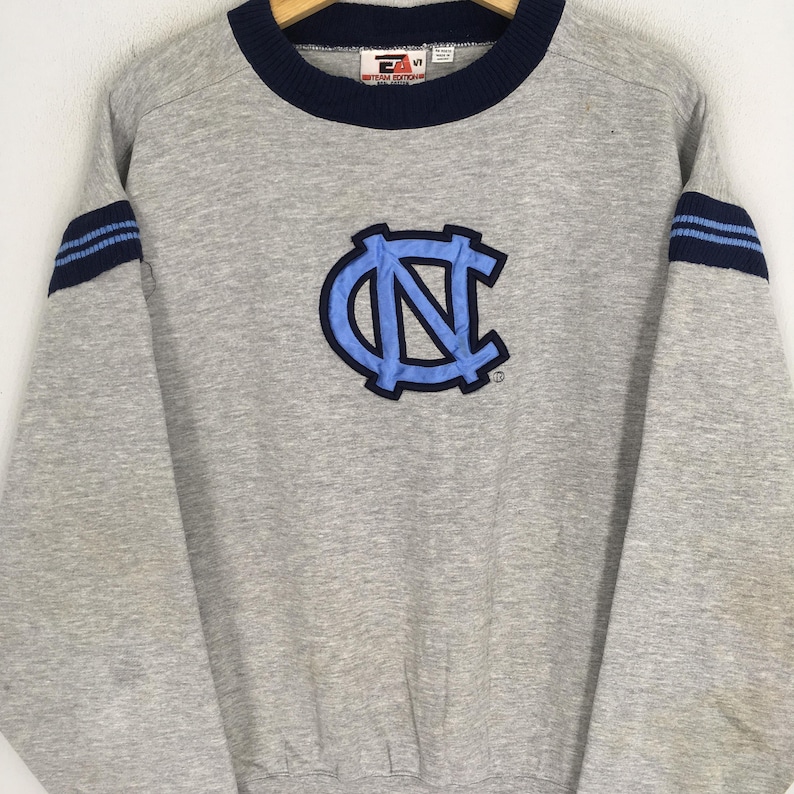 K&ouml;nnte beinhalten: Ein graues Sweatshirt mit marineblauem Kragen und &Auml;rmelstreifen. Auf der Vorderseite befindet sich ein hellblaues und marineblaues Logo. Das Sweatshirt hat lange &Auml;rmel und einen gerippten Saum. Das Logo ist ein stilisiertes "NC"-Design.