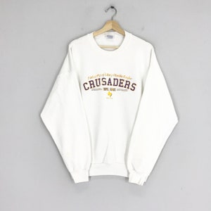 Op de afbeelding: Witte trui met een paarse en gouden afbeelding met de tekst "University of Mary Hardin-Baylor Crusaders Est. 1893" met een kleine omtrek van de staat Texas.