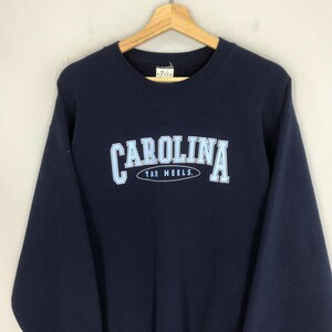 May include: Navy blue sweatshirt with the word "CAROLINA" in light blue block letters, arched above "TAR HEELS." The sweatshirt is hanging on a wooden hanger against a neutral background. The sweatshirt is a crew neck style.