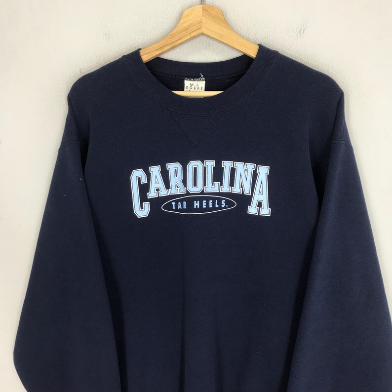 May include: Navy blue sweatshirt with the word "CAROLINA" in light blue block letters, arched above "TAR HEELS." The sweatshirt is hanging on a wooden hanger against a neutral background. The sweatshirt is a crew neck style.