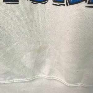 May include: White sweatshirt with a blue and black graphic print. The sweatshirt has a slight discoloration on the front.