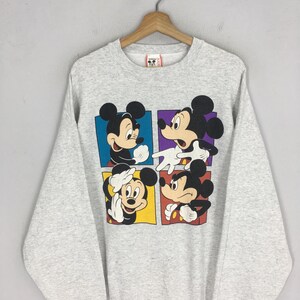 May include: A light gray sweatshirt featuring a colorful graphic of Mickey Mouse in four different poses. Each Mickey Mouse is in a different colored square. The sweatshirt is hanging on a wooden hanger.