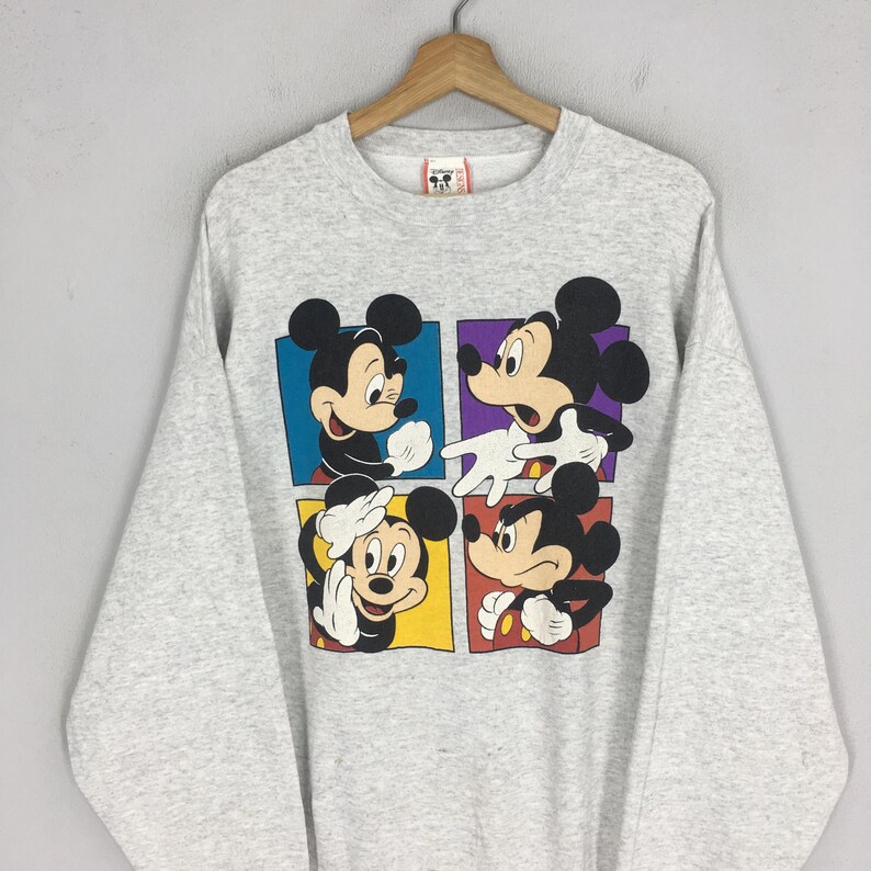 May include: A light gray sweatshirt featuring a colorful graphic of Mickey Mouse in four different poses. Each Mickey Mouse is in a different colored square. The sweatshirt is hanging on a wooden hanger.