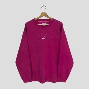 Könnte beinhalten: Ein magentafarbenes Sweatshirt mit V-Ausschnitt und langen Ärmeln. Der gestickte Text "Cape Cod" ist über der Brust gewölbt, mit einer weißen Umrisslinie einer Walflosse. Der Text "Massachusetts" steht darunter.