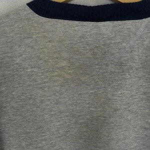 K&ouml;nnte beinhalten: Ein graues Sweatshirt mit Rundhalsausschnitt und marineblauem Kragen. Der Stoff scheint ein weiches Strickmaterial zu sein. Das Sweatshirt hat Verf&auml;rbungen auf der Vorderseite.